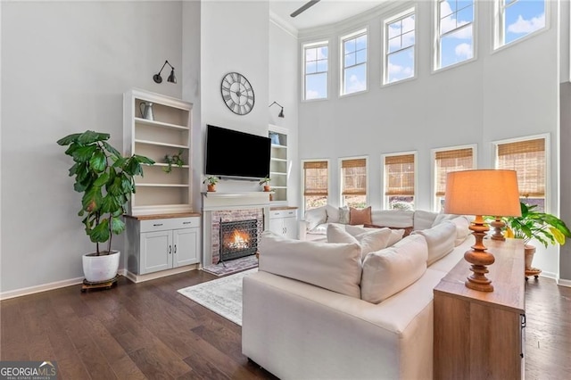 living room with a warm lit fireplace, a towering ceiling, baseboards, and dark wood-style flooring