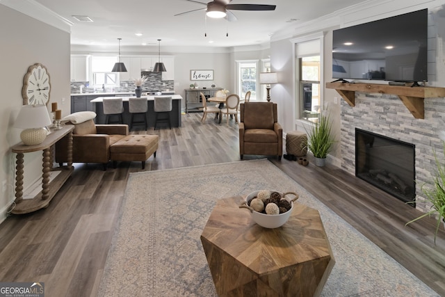 living room with crown molding, wood finished floors, a stone fireplace, ceiling fan, and recessed lighting