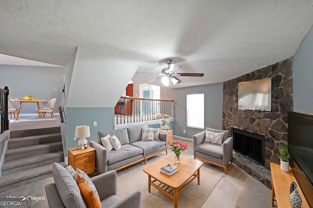 living area featuring wood finished floors, ceiling fan, a stone fireplace, a textured ceiling, and baseboards