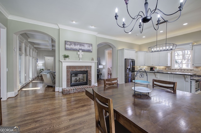 dining space featuring dark wood-type flooring, ornamental molding, a chandelier, and a brick fireplace