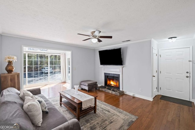 living room featuring ornamental molding, a ceiling fan, a textured ceiling, dark wood finished floors, and a stone fireplace