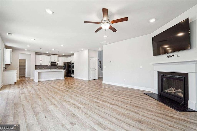 unfurnished living room featuring a sink, a glass covered fireplace, light wood finished floors, baseboards, and ceiling fan