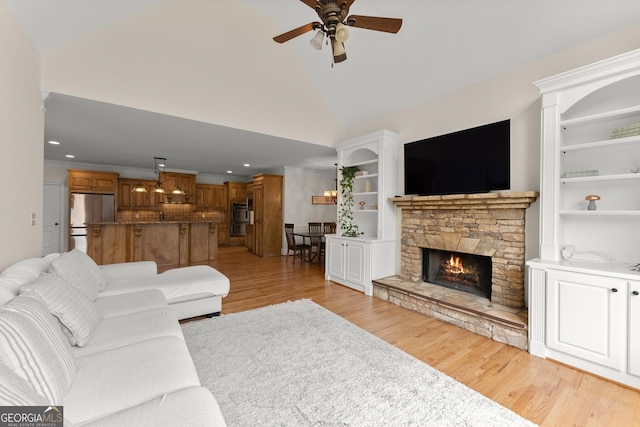 living area featuring built in features, high vaulted ceiling, ceiling fan, a stone fireplace, and light wood-style floors