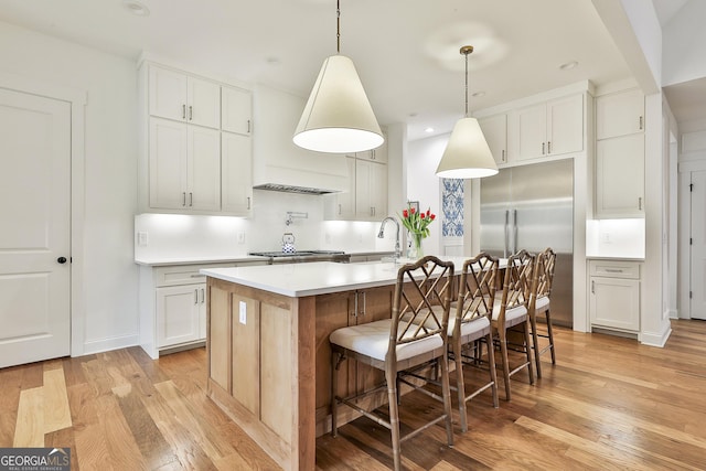 kitchen with light countertops, stainless steel built in refrigerator, a center island with sink, white cabinets, and light wood-style floors