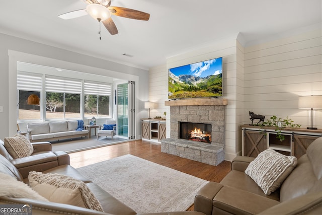 living room with wood finished floors, a ceiling fan, a stone fireplace, wooden walls, and ornamental molding