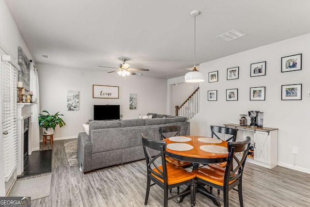 dining room with stairs, a fireplace with raised hearth, light wood-style flooring, and a ceiling fan