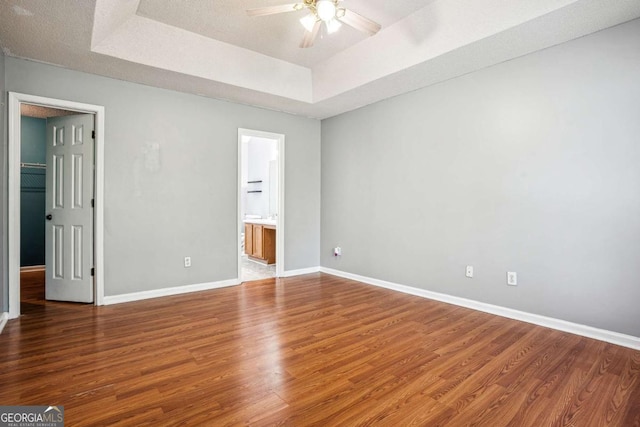 unfurnished bedroom featuring a tray ceiling, a textured ceiling, ensuite bath, wood finished floors, and baseboards