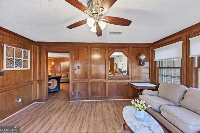 living area with a ceiling fan, wooden walls, a textured ceiling, visible vents, and light wood-style floors