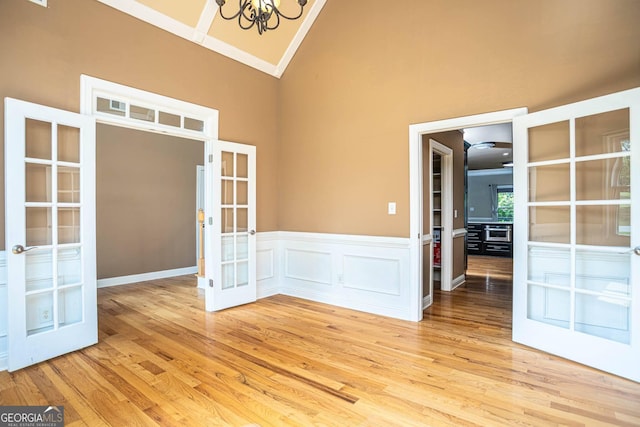 unfurnished room with a notable chandelier, french doors, a wainscoted wall, light wood-style floors, and high vaulted ceiling
