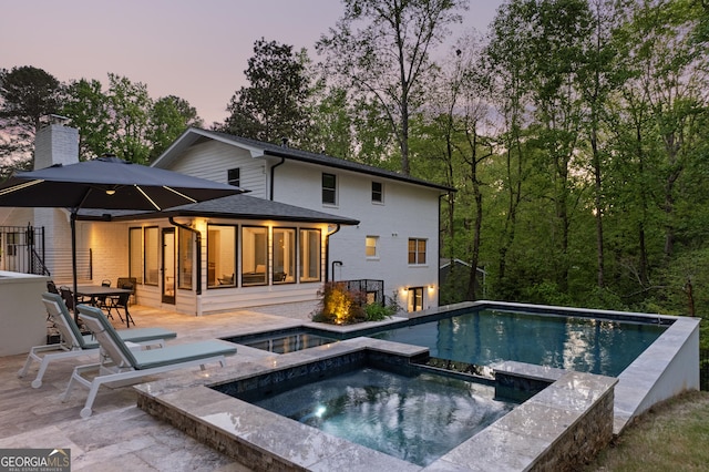 back of property at dusk featuring a chimney, a patio area, and a pool with connected hot tub