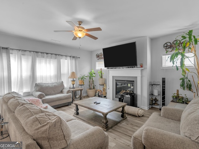 living room with light wood-type flooring, visible vents, a glass covered fireplace, and a ceiling fan