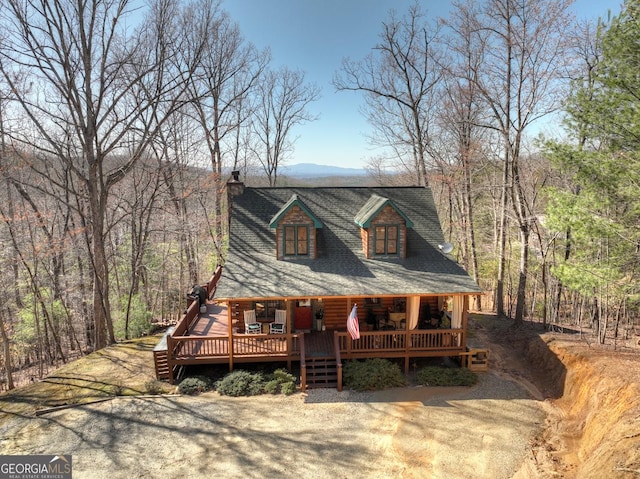 view of front of home with a chimney, a wooded view, covered porch, and a shingled roof