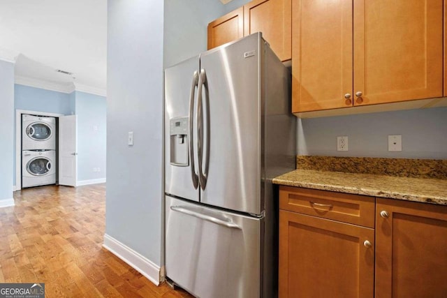 kitchen with stainless steel fridge, ornamental molding, light stone counters, stacked washer / dryer, and light wood-style flooring