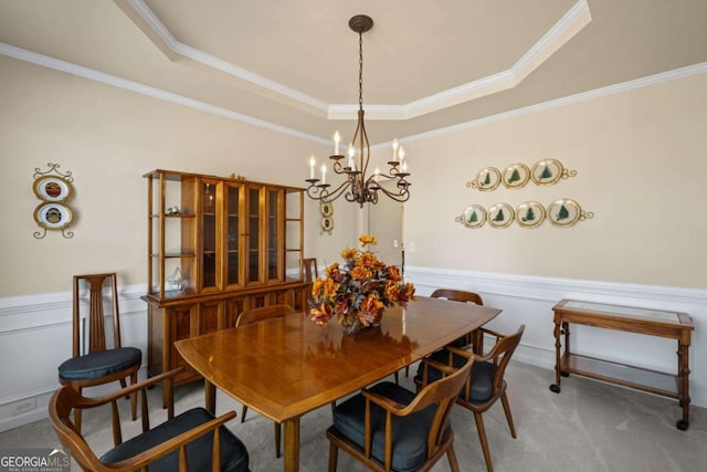 carpeted dining space with an inviting chandelier, a raised ceiling, ornamental molding, and wainscoting