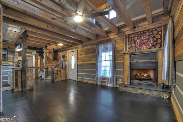 unfurnished living room with a ceiling fan, stairway, beamed ceiling, wooden walls, and a warm lit fireplace