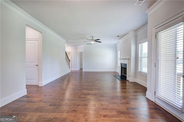 unfurnished living room with a fireplace with raised hearth, dark wood-type flooring, crown molding, stairs, and ceiling fan