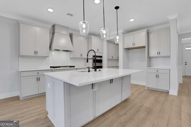 kitchen featuring custom range hood, a center island with sink, a sink, light wood-style flooring, and stovetop