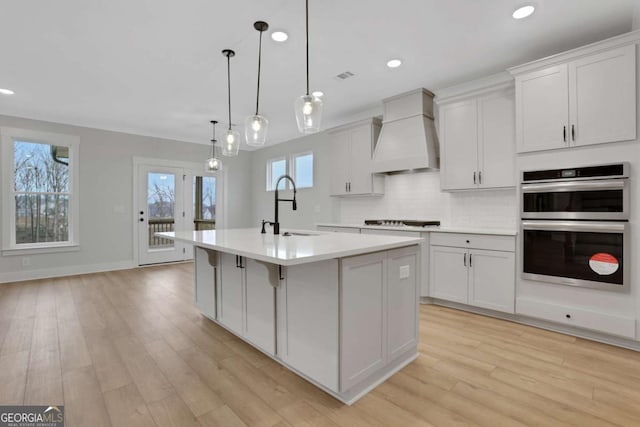 kitchen with an island with sink, a sink, tasteful backsplash, stainless steel double oven, and custom exhaust hood