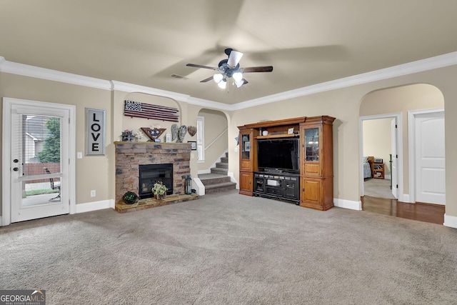 carpeted living room with crown molding, baseboards, arched walkways, ceiling fan, and a stone fireplace
