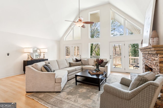 living area featuring high vaulted ceiling, a ceiling fan, wood finished floors, a stone fireplace, and baseboards