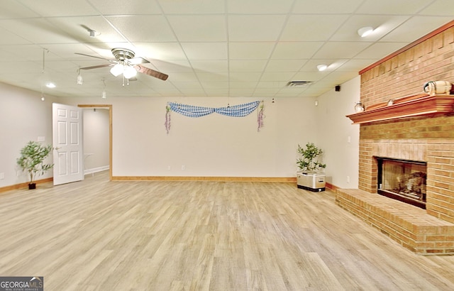 unfurnished living room featuring wood finished floors, a drop ceiling, ceiling fan, a brick fireplace, and baseboards