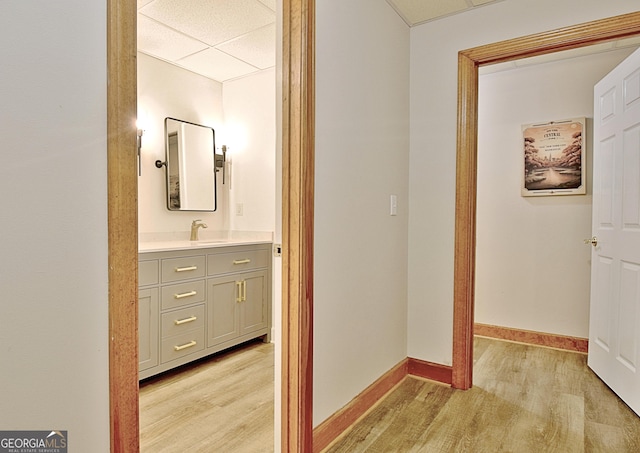 hall with light wood finished floors, a sink, a drop ceiling, and baseboards