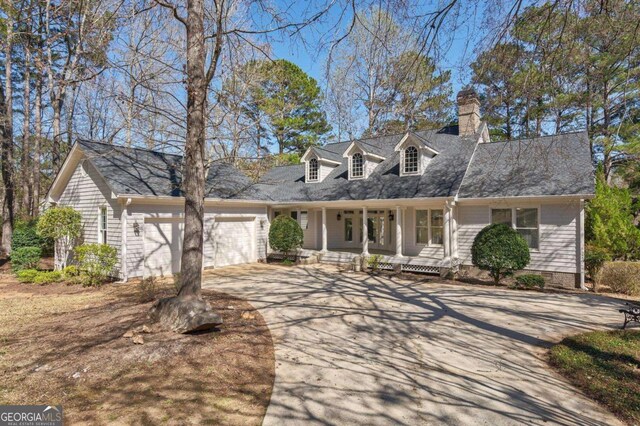 new england style home with driveway, covered porch, an attached garage, and a chimney