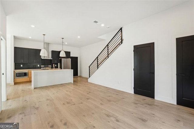 kitchen with tasteful backsplash, hanging light fixtures, stainless steel appliances, a kitchen island with sink, and light wood-style floors