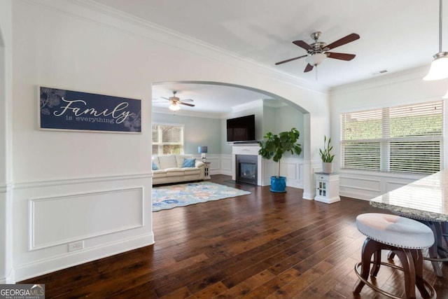living area featuring crown molding, a decorative wall, a ceiling fan, visible vents, and a glass covered fireplace