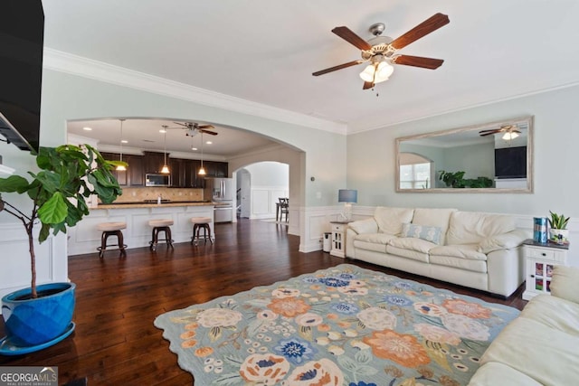 living room with crown molding, arched walkways, a ceiling fan, a wainscoted wall, and dark wood-style flooring