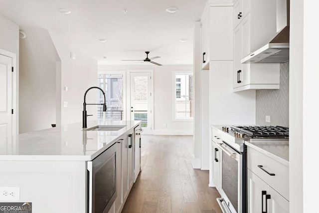 kitchen featuring gas range, a sink, wall chimney exhaust hood, ceiling fan, and light wood-style flooring