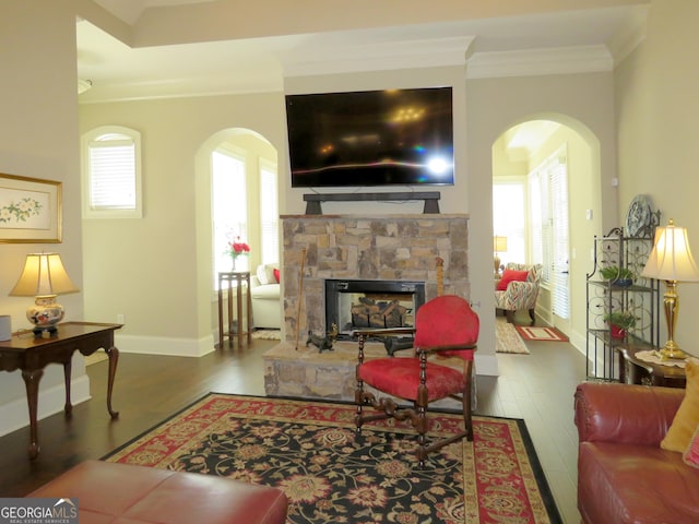 living room featuring ornamental molding, arched walkways, a stone fireplace, and wood finished floors