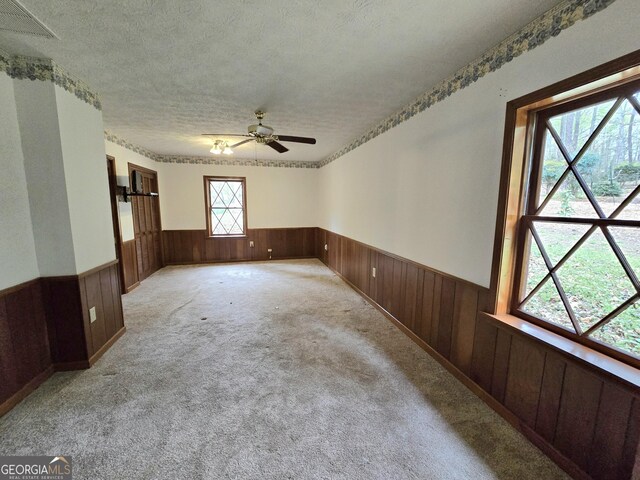 empty room featuring a textured ceiling, wood walls, carpet, ceiling fan, and wainscoting