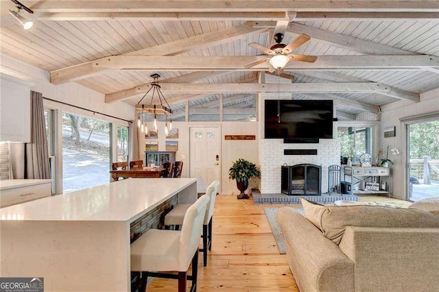 living room with a brick fireplace, light wood-style floors, ceiling fan with notable chandelier, vaulted ceiling with beams, and wooden ceiling