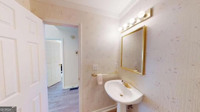 bathroom featuring wood finished floors, wallpapered walls, ornamental molding, a sink, and baseboards
