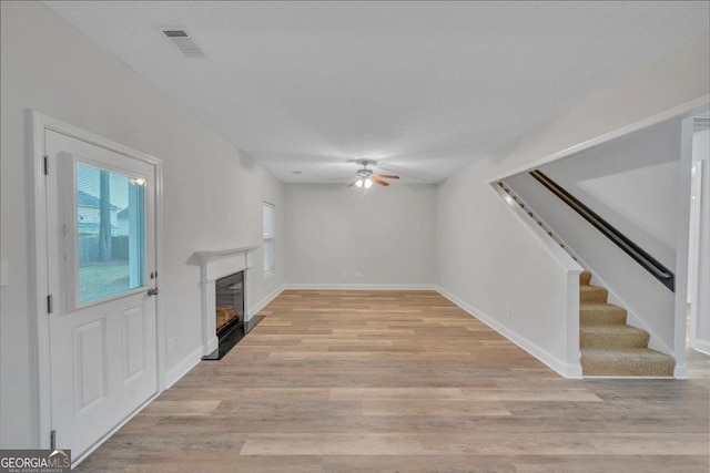 unfurnished living room featuring stairway, light wood finished floors, ceiling fan, visible vents, and a glass covered fireplace