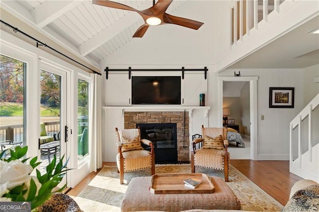 living area with a stone fireplace, light wood-type flooring, a ceiling fan, beamed ceiling, and high vaulted ceiling