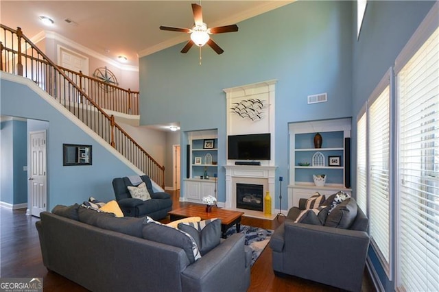 living room featuring ceiling fan, a towering ceiling, a glass covered fireplace, visible vents, and stairway