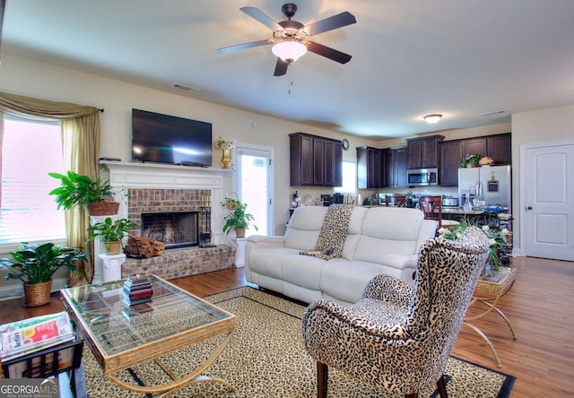 living room featuring a brick fireplace, ceiling fan, visible vents, and light wood finished floors