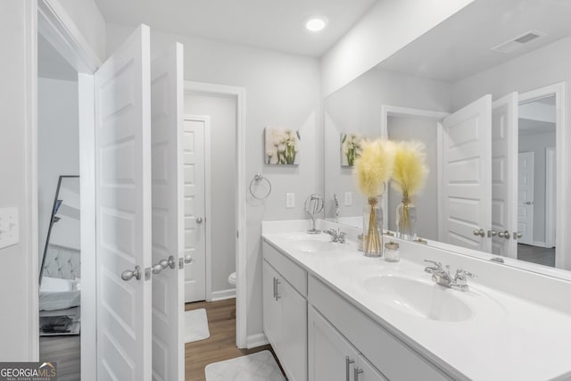 ensuite bathroom featuring a sink, double vanity, toilet, wood finished floors, and visible vents