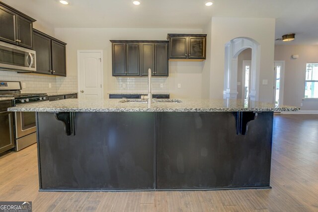 kitchen featuring appliances with stainless steel finishes, arched walkways, a kitchen island with sink, and light stone counters