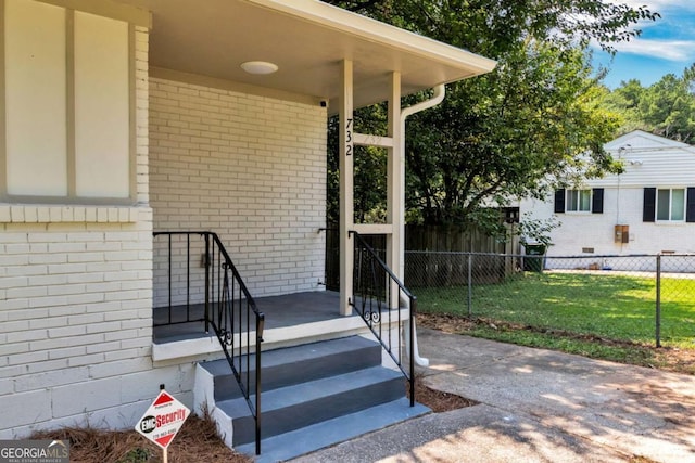 view of exterior entry with a lawn, brick siding, and fence
