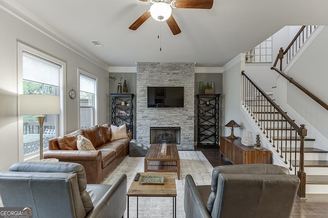 living area with wood finished floors, a ceiling fan, ornamental molding, a stone fireplace, and stairs