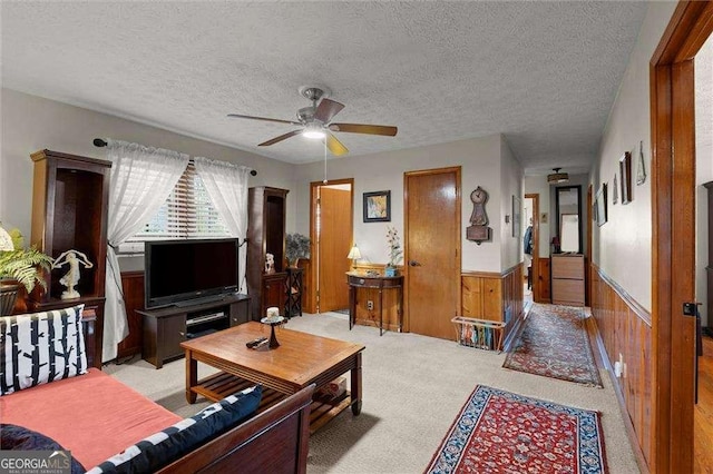 living area with ceiling fan, wooden walls, light colored carpet, a textured ceiling, and a wainscoted wall
