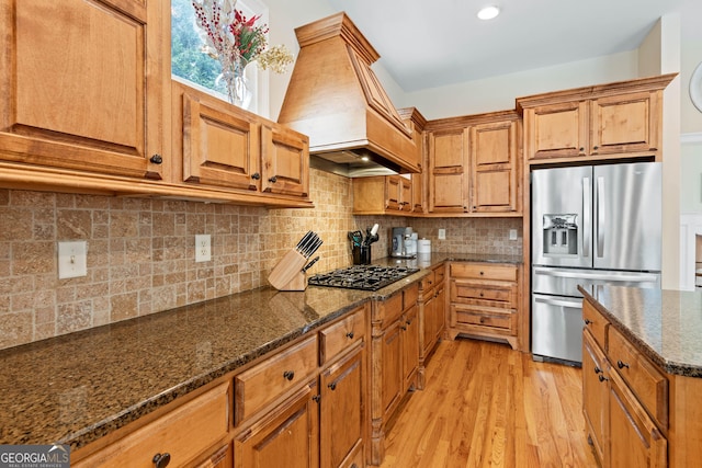 kitchen featuring custom exhaust hood, gas cooktop, stainless steel refrigerator with ice dispenser, brown cabinetry, and light wood finished floors
