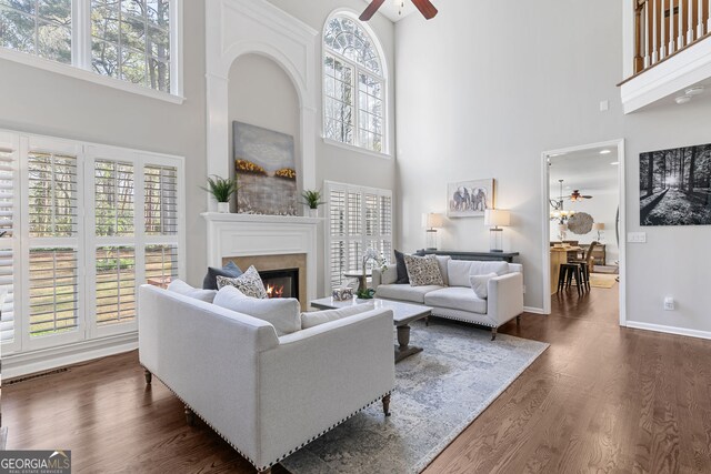 living area with a ceiling fan, dark wood-style flooring, baseboards, a high ceiling, and a warm lit fireplace