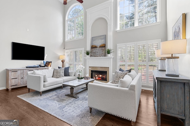 living room with dark wood-style flooring, baseboards, a towering ceiling, ceiling fan, and a glass covered fireplace