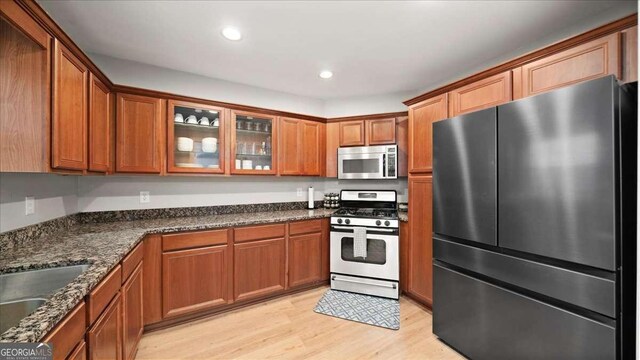 kitchen featuring stainless steel appliances, glass insert cabinets, dark stone countertops, light wood-style floors, and brown cabinetry