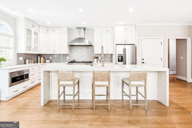 kitchen featuring decorative backsplash, light countertops, light wood-style flooring, stainless steel appliances, and wall chimney range hood