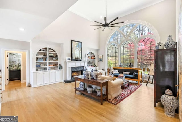 living area with a warm lit fireplace, built in features, ceiling fan, and light wood-type flooring